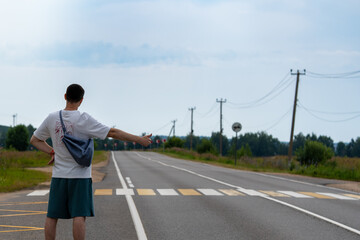 Man with a bag hitchhikes on a rural road by extending his thumb. The wide road stretches into the distance with a vehicle approaching, highlighting travel and adventure themes. © magpie