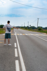 Man with a bag hitchhikes on a rural road by extending his thumb. The wide road stretches into the distance with a vehicle approaching, highlighting travel and adventure themes. © magpie