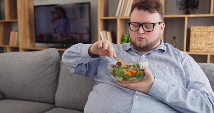 Overweight man eating salad at home watching tv. On the sofa he watches cooking show and eats greens, choosing light meals for weight loss. Healthy diet wellness and lifestyle concept.