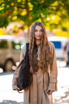 Young woman with long wavy brown hair in tan trench coat and high waist skirt carrying shoulder bag outdoors in soft sunlight with warm blurred greenery behind, with woman, portrait, hair, longhair