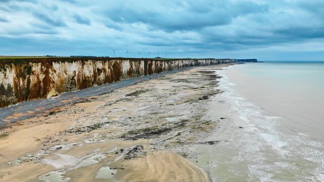 Normandy chalk cliff coastline above wide low tide beach on English Channel, France. Coastal travel landscape with wind turbines background
