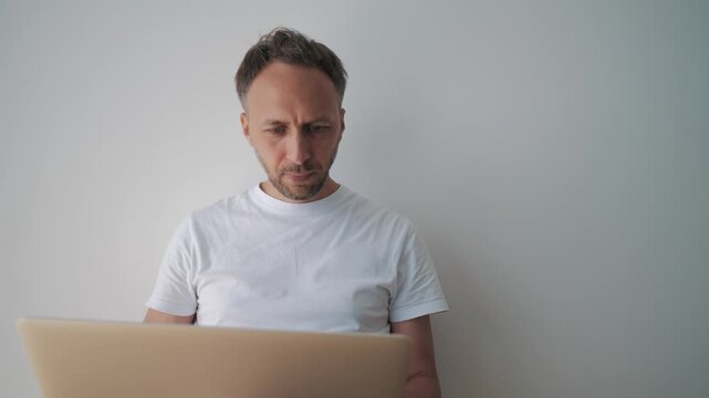 Concerned professional reads laptop screen against plain wall, conveying digital overload, financial anxiety, job uncertainty, or online scam awareness.