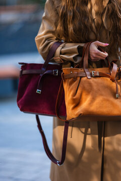 Woman carrying suede tote and burgundy shoulder bag, wearing beige trench coat with long wavy hair, pink manicure, and stylish urban fashion details in closeup, with bag, tote, suede, handbag detail