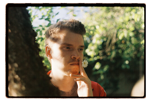 A young man holds a daisy to his lips among summer trees