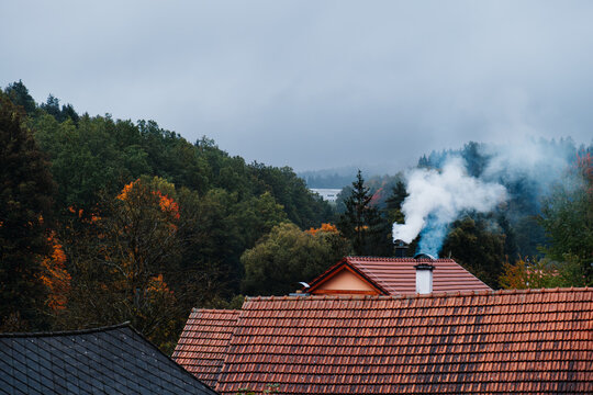Misty autumn village rooftops with chimney smoke, colorful forest trees and foggy hills, quiet rural landscape on an overcast rainy day