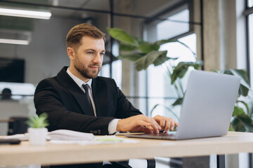 Fototapeta premium Businessman in a suit working diligently on a laptop at a desk in a contemporary open plan office setting