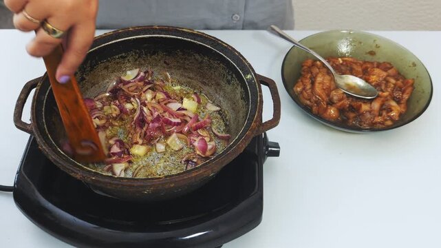Onions are fried in vegetable oil in a deep cast-iron skillet and stirred with a spatula. Medium shot.