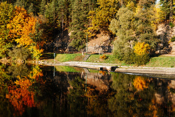 Autumn riverside forest with colorful trees reflected in calm water, stone shore © Mariana Verbenets