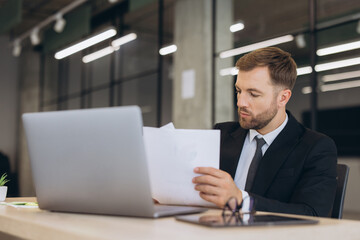 Fototapeta premium Businessman analyzing documents and data on laptop, working at office desk and planning strategy