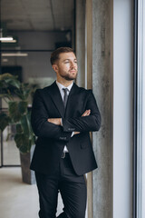 Fototapeta premium Confident businessman in suit with folded arms standing by an office window looking into the future
