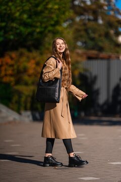 Young woman in tan trench coat smiling on sunny city street with black leather tote, casual chic autumn fashion, confident pose, soft daylight, urban backdrop, with woman, trenchcoat, tan, coat detail