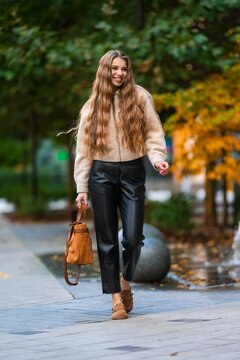 Autumn street style portrait of woman walking outdoors in beige zip jacket, black leather pants, and tan boots along tree lined park path with colorful foliage, with woman, autumn, fall, outdoors