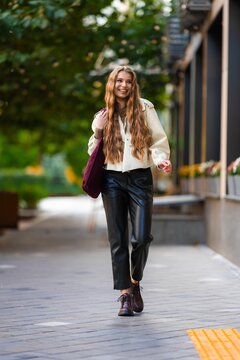 Smiling young woman walking along urban sidewalk in beige jacket, black leather pants, and burgundy bag beside greenery and modern city buildings, with woman, young, smiling, walking detail
