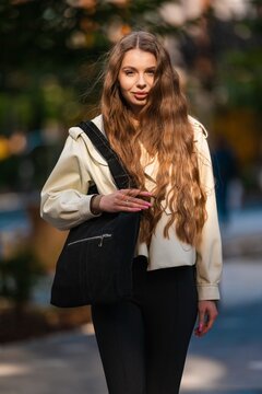 Portrait of young woman outdoors with long wavy hair, cream jacket, black tote bag, soft smile, and warm sunlit urban park setting with blurred background, with woman, portrait, outdoor, lighting
