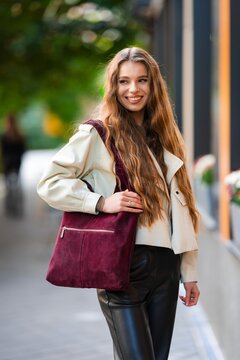 Smiling young woman on sunny city sidewalk wearing cream jacket and burgundy suede tote bag, with long wavy hair, casual urban fashion style, relaxed daytime portrait