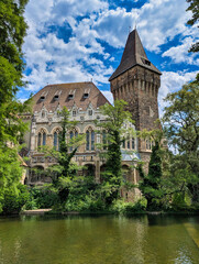 Obraz premium Gothic and Romanesque sections of Vajdahunyad Castle reflected in a surrounding moat, with lush vegetation and historic stone architecture, City Park, Budapest, Hungary.