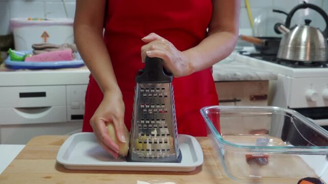 A woman in a red apron holds a grater and a peeled potato. She prepares to grate vegetables with calm focus and care. She enjoys making a fresh meal in her cozy kitchen.