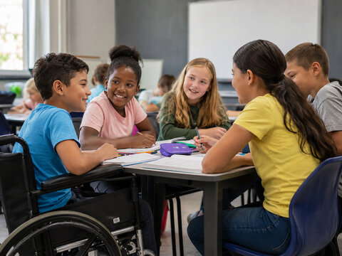 Inclusive group of elementary students, including a child using a wheelchair, collaborate around a classroom table with notebooks and pencils.