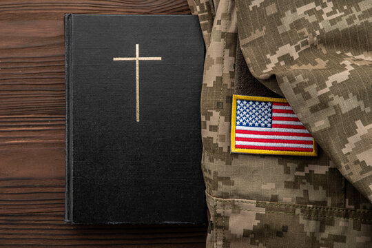 Christian cross on black book beside military camouflage uniform with American flag US, concept of faith and military service