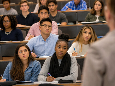 Diverse university students listen attentively and take notes in a tiered lecture hall during a college class.