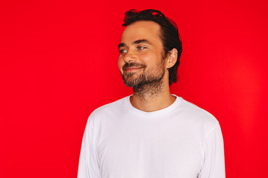 Studio Portrait of a Young Caucasian Man with Dark Hair and Beard Wearing White Long-Sleeve T-Shirt Looking Away with a Sly, Knowing, and Slight Smiling Expression Isolated on Solid Red Background