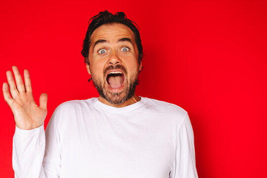 Studio portrait of a surprised shocked young caucasian man with dark hair and beard wearing white long sleeve t shirt wide eyes open mouth waving hand gesturing greeting or shock on red background
