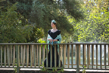 Fototapeta premium A young woman from Andalusia wearing a black flamenco dress with ruffles and an aquamarine shawl, with flowers in her hair, posing on a wooden bridge in an outdoor park. Concept: dance, art.