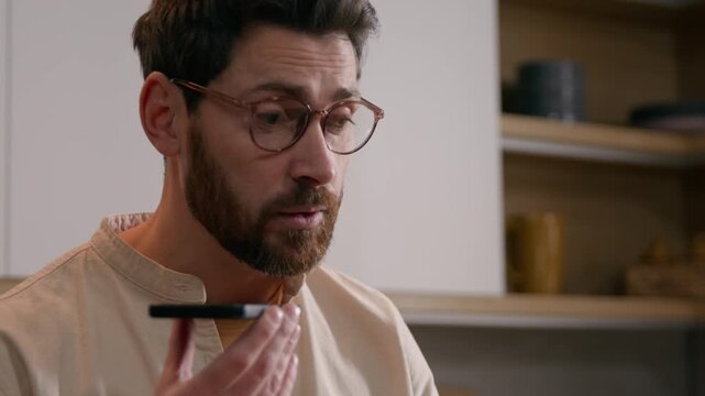 Caucasian man recording voice message on smartphone talking mobile phone using voicemail at kitchen remote freelancer businessman communicating cellphone app focused male speaking business at home