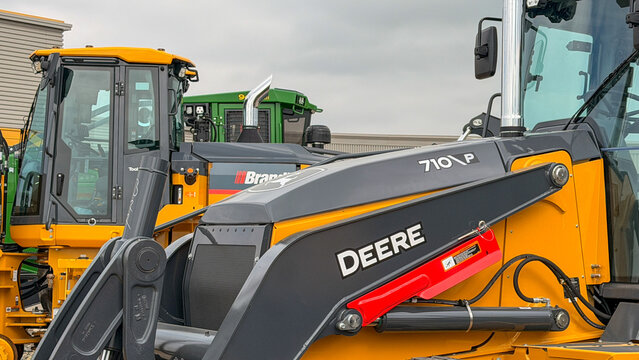 Regina, Canada - July 20, 2026: John Deere 710P backhoe loader at Brandt dealership lot with green tractor visible in background