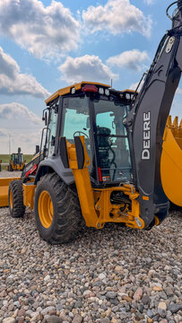 Regina, Canada - July 20, 2026: John Deere yellow backhoe loader parked on gravel lot with cab detail and boom arm visible under partly cloudy sky