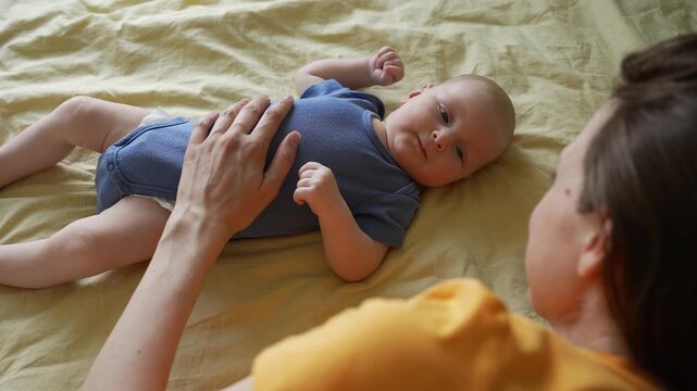 Young loving mother tenderly stroking her newborn baby son's chest while he lies on the bed. Caring mom and her infant child sharing an intimate bonding moment in the bedroom at home