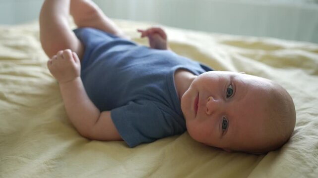 Sweet infant with bright blue eyes wearing a blue bodysuit and lying on his back on a comfortable bed. The curious newborn is actively moving his limbs, looking directly at the camera