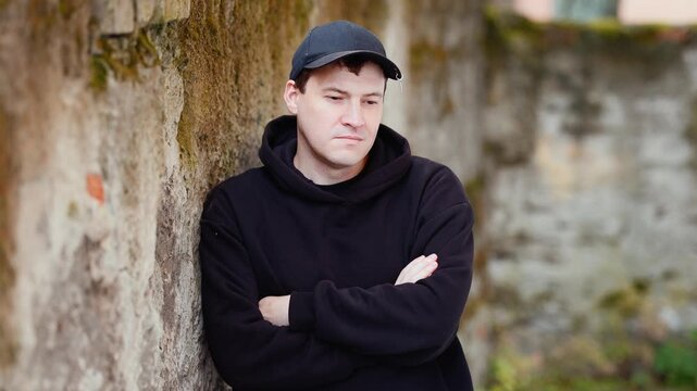 Man in black hoodie and cap leaning against rustic stone wall. Casual portrait. Urban street style outdoor session.