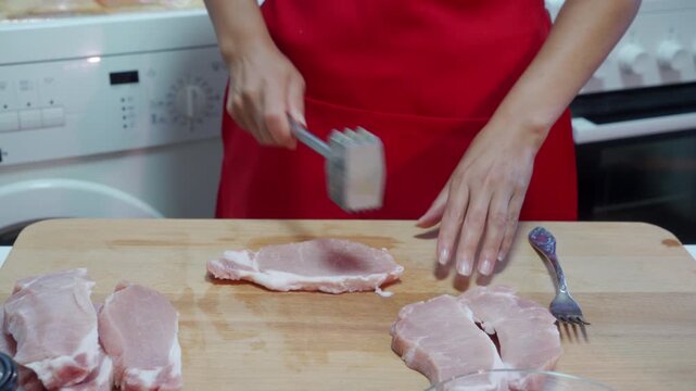 A determined woman uses a meat mallet to pound raw pork chops on a wooden cutting board in a kitchen. She feels confident and focused while preparing a delicious dinner.