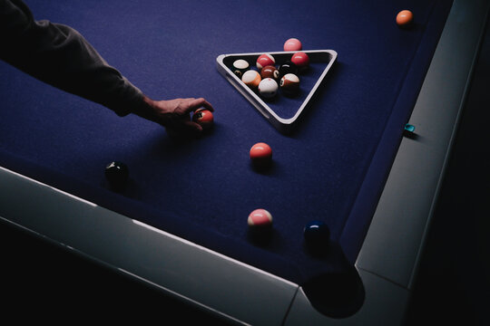 Player&rsquo;s hand positions a billiard ball beside a triangle rack on a dark blue pool table in a dimly lit game room