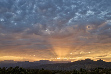 Fototapeta premium Dramatic sunrise over the Mojave desert mountains near Joshua Tree, with golden sunbeams breaking through blue and orange clouds.