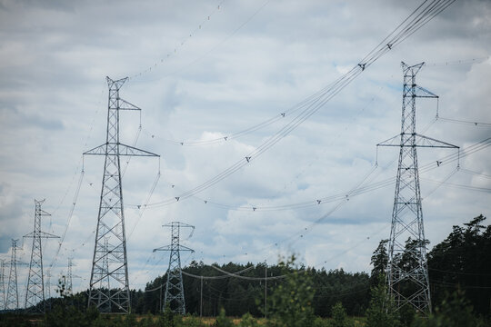 High voltage power lines and transmission towers against a cloudy sky, surrounded by green vegetation and trees in a rural landscape