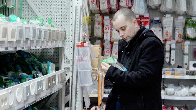 Electrician examining light switch specifications in hardware store, surrounded by electrical tools and components, verifying technical details before installation