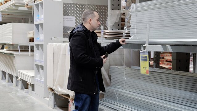 Construction worker examining metal profiles, comparing specifications on smartphone while standing in warehouse, selecting materials for upcoming renovation project