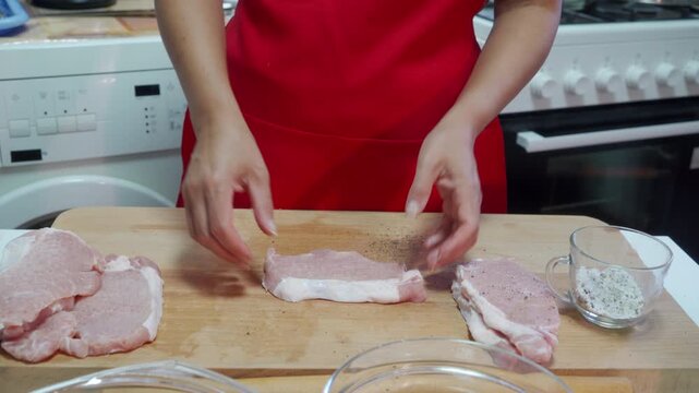 cook carefully seasons pork chops in kitchen, using salt shaker on wooden cutting board, stove in background, red apron, white and brown colors, focused overhead view, commercial food prep.
