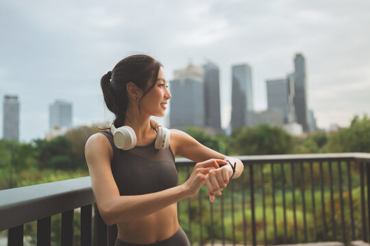Fit Asian woman standing at city park bridge looking away while checking smartwatch after running workout, active sporty girl wearing wireless headphones health tracking and urban fitness lifestyle
