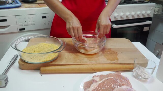 A person in a red apron dips raw pork into a bowl of egg wash. The kitchen atmosphere feels calm and purposeful. Preparations bring joy as the meal is crafted with care.