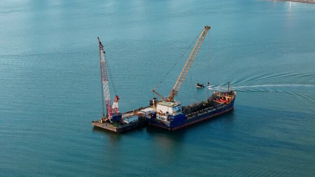 Coastal dredging vessel operating with floating crane platform in open water. Aerial view of seabed material handling and marine engineering work