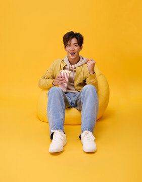 Movie entertainment concept with excited Asian man holding popcorn and cheering on beanbag in studio expressing leisure streaming enjoyment positive emotion and youthful home cinema lifestyle.