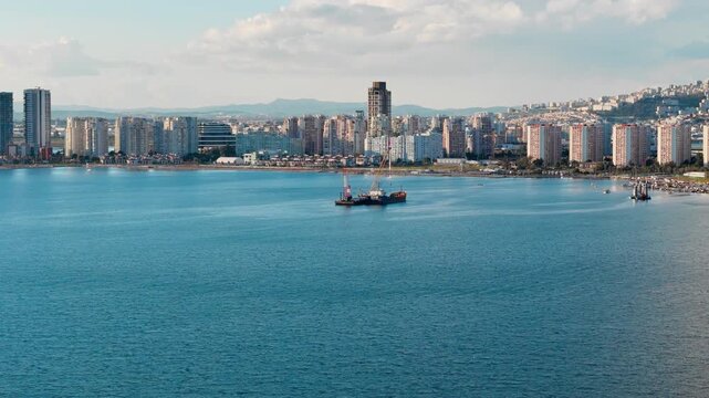 Wide aerial view of dredging operation in Izmir Bay with modern residential skyline along the shore. Marine maintenance vessels positioned in calm urban coastal water