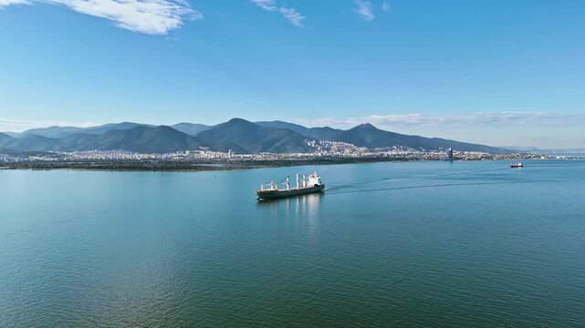 Dry bulk cargo ship sailing through calm sea near coastal city. Aerial view for global trade, import export logistics and maritime freight transport
