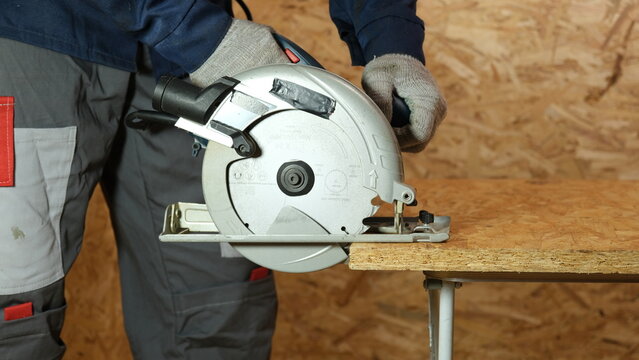 Builder saws a board with a circular saw cutting a wooden plank in a joinery. Carpenter uses portable circular saw tool to cut wood at wooden table in workshop. Worker sawing pine board. Real image