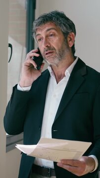 Video of elegant businessman reading some notes while talking with smartphone standing  at the office.
