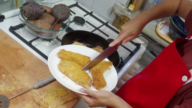 A caring cook places crisp fried cutlets onto a white plate. She uses a knife near the warm stove in a cozy home kitchen. Serving this comforting food brings deep joy.