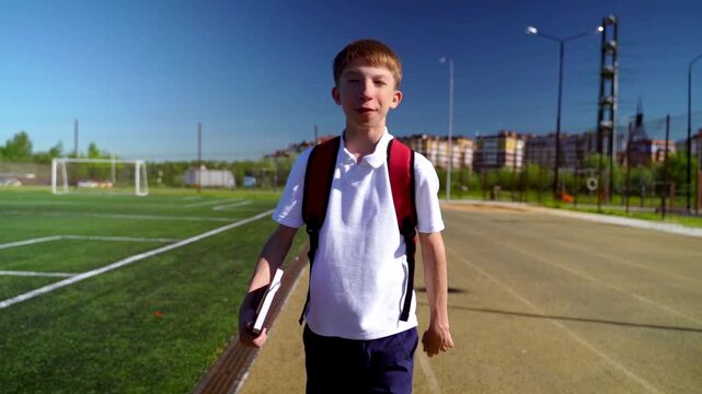 Young boy walks toward camera on sunny day. He carries backpack and school book. He smiles slightly, looking calm and cheerful. Sidewalk runs beside green soccer field